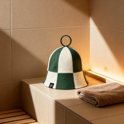 Koala Hats green and natural checkered sauna hat made from 100% Australian merino wool, placed on a tiled sauna bench beside a folded towel, designed to regulate head temperature, protect hair and scalp, and help users stay in the sauna longer with greater comfort.