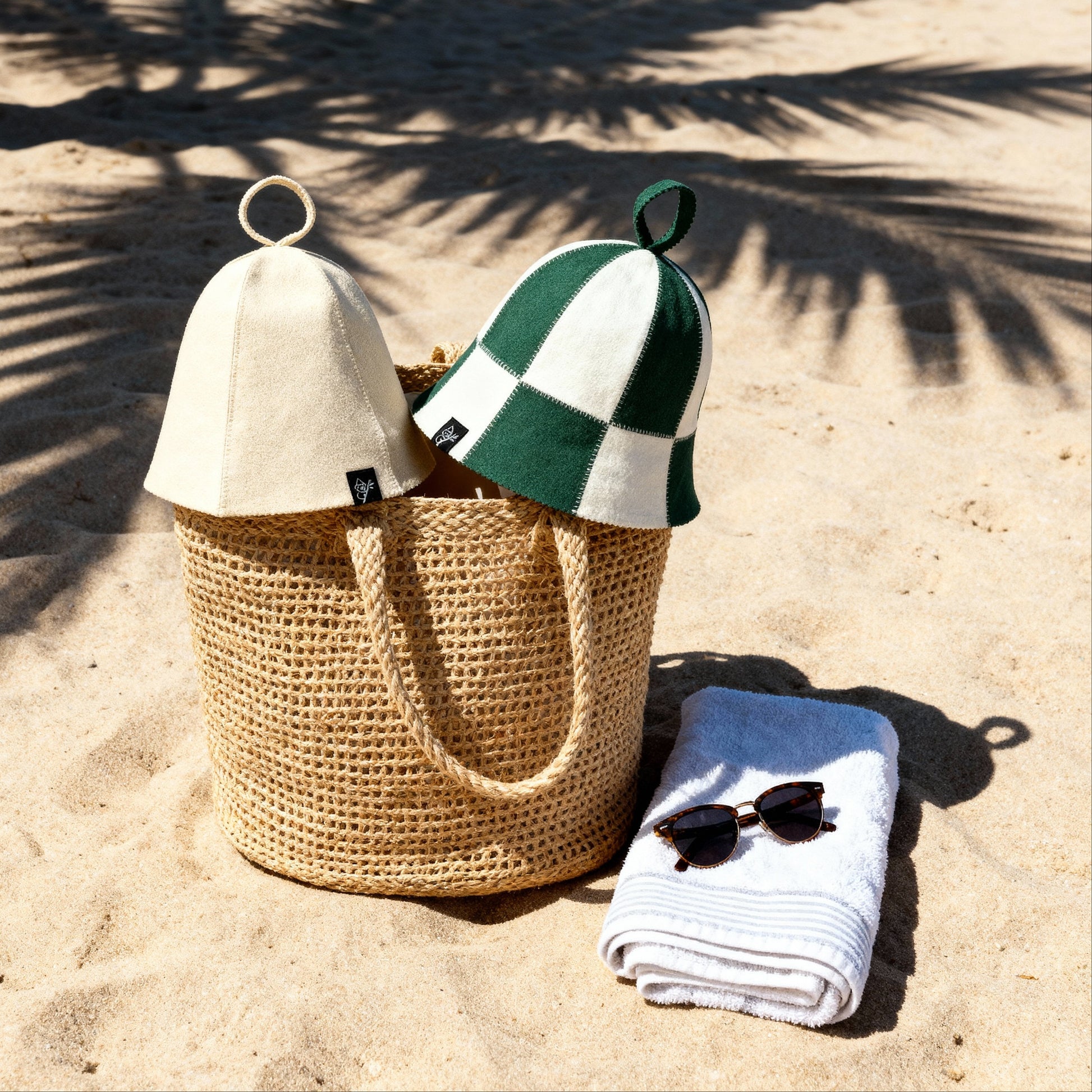 Two Koala Hats premium sauna hats made from Australian merino wool, including a natural and green checkered design, displayed in a woven beach bag on sand, highlighting Koala Hats as a lifestyle sauna accessory for heat regulation, scalp protection, and longer sauna sessions.