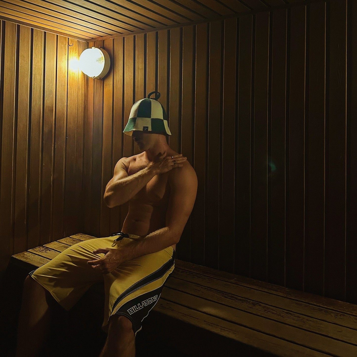Man sitting on a wooden sauna bench wearing a Koala merino wool sauna hat, using the hat to regulate head temperature, reduce heat stress on the scalp, protect hair, and extend sauna sessions for improved recovery and overall sauna performance.