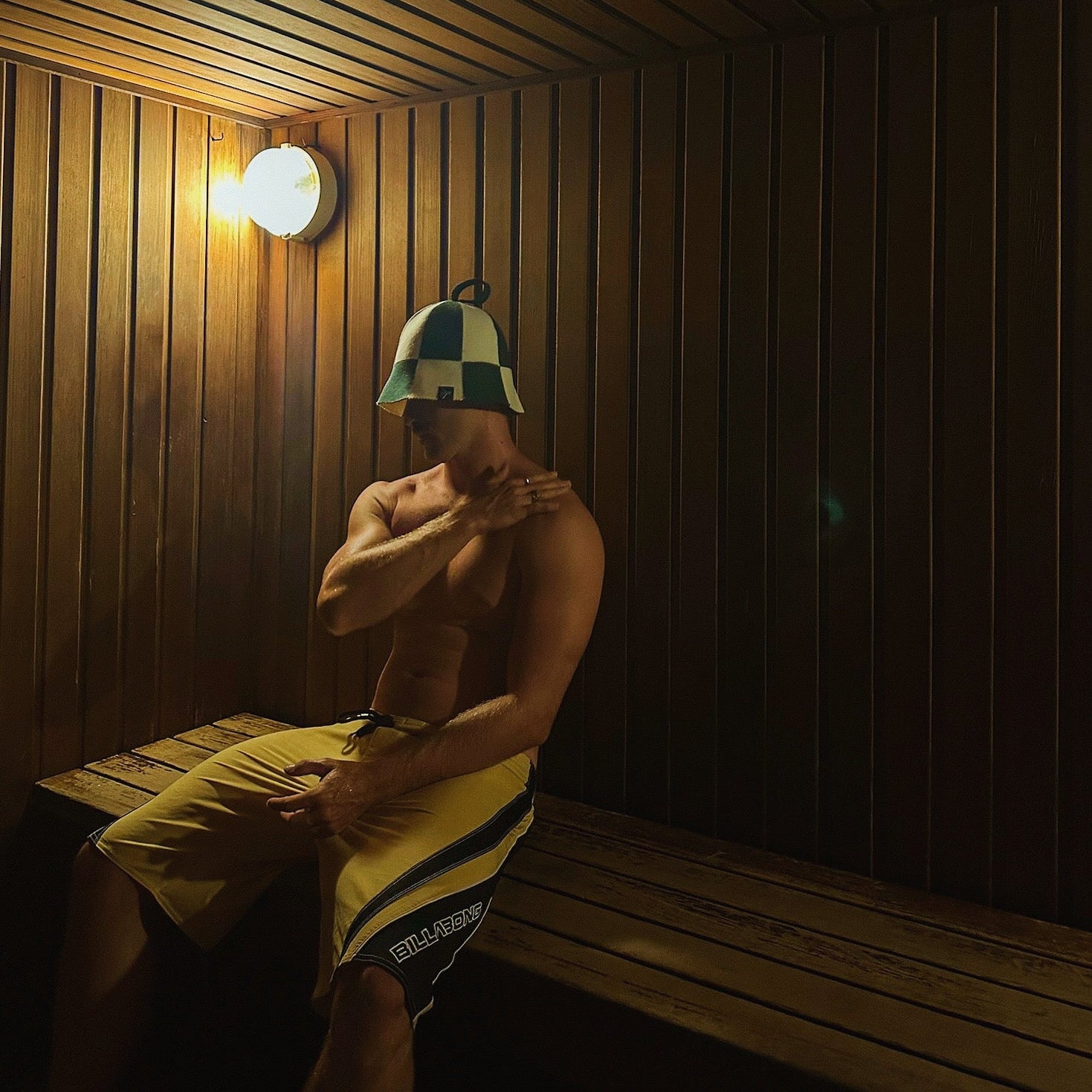 Man sitting on a wooden sauna bench wearing a Koala merino wool sauna hat, using the hat to regulate head temperature, reduce heat stress on the scalp, protect hair, and extend sauna sessions for improved recovery and overall sauna performance.